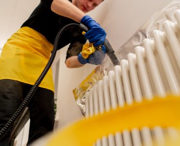 low shot in an apartment a cleaner in a yellow apron washes a white radiator with a steam generator