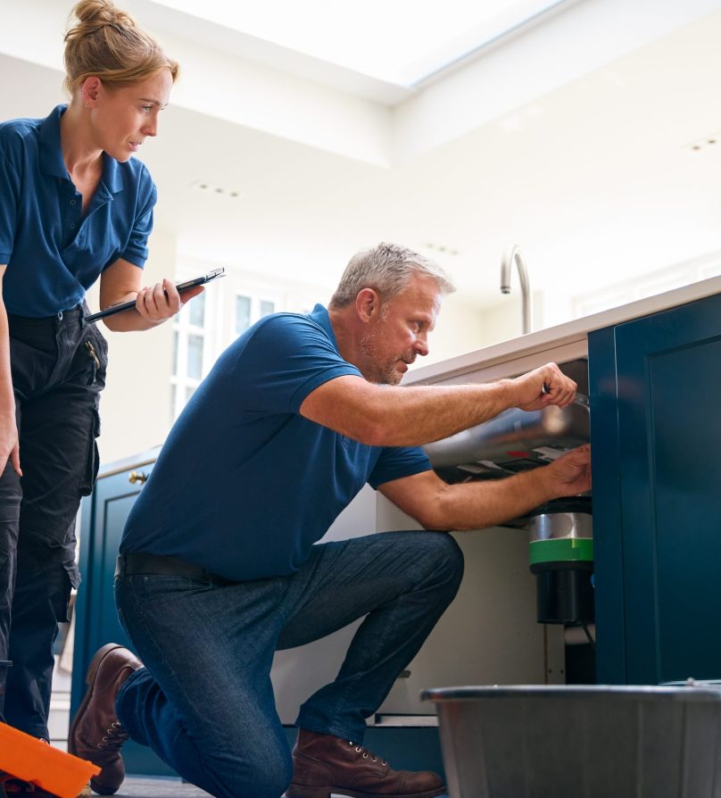 Male Plumber With Female Trainee Using Digital Tablet Fixing Waste Disposal Unit In Kitchen Sink