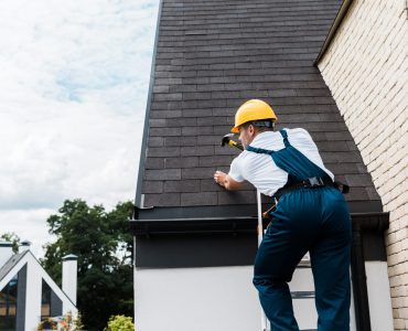 repairman in uniform and helmet repairing roof while standing on ladder