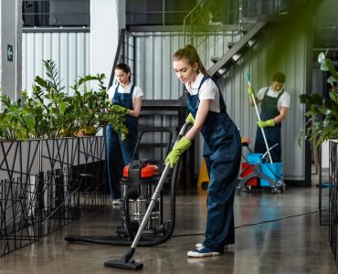 young cleaner vacuuming floor in office near colleagues