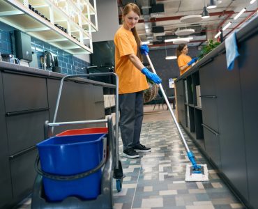 Young woman cleaning the floor with a mop, her colleague is in the background